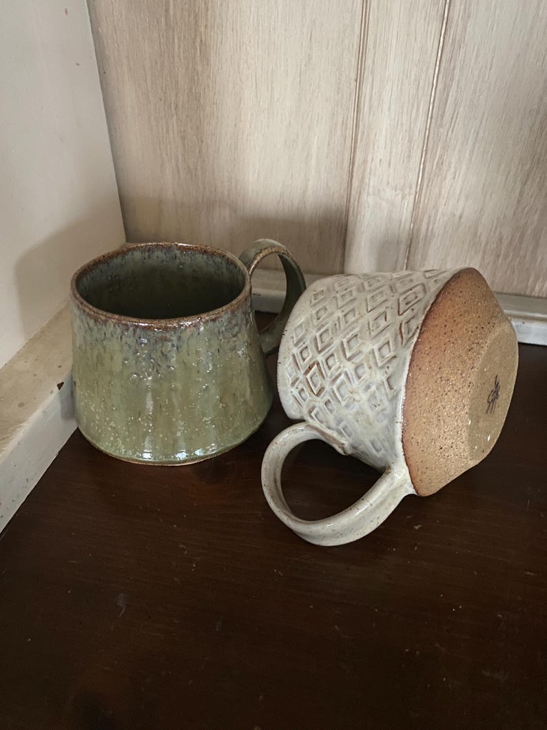 May include: Two ceramic mugs. One mug is upright with a mottled olive green glaze and a smooth finish. The second mug is lying on its side, featuring a cream-colored exterior with a raised geometric diamond pattern and a textured brown base.