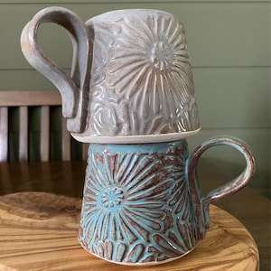 May include: Two stacked ceramic mugs featuring a raised, textured floral pattern. The top mug is a muted lavender-gray, while the bottom mug is a vibrant turquoise blue with brown undertones. Each mug has a large, curved handle and a wide base.