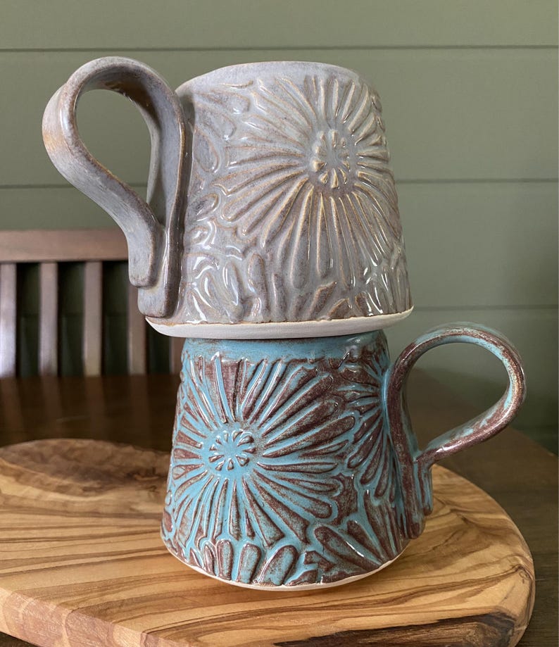 May include: Two stacked ceramic mugs featuring a raised, textured floral pattern. The top mug is a muted lavender-gray, while the bottom mug is a vibrant turquoise blue with brown undertones. Each mug has a large, curved handle and a wide base.