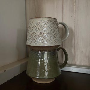 May include: Two stacked ceramic mugs with handles. The top mug is off-white with a raised geometric diamond pattern and a brown base. The bottom mug is a glossy olive green with a textured, speckled finish and a wide, flat base.