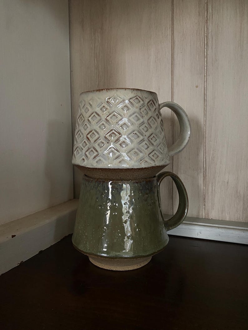 May include: Two stacked ceramic mugs with handles. The top mug is off-white with a raised geometric diamond pattern and a brown base. The bottom mug is a glossy olive green with a textured, speckled finish and a wide, flat base.
