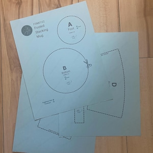 May include: Light blue paper templates for a footed stacking mug, labeled FSM01V2. The sheets feature printed diagrams with dashed cutting lines, scissors icons, and labels including A Foot, B Bottom, and D. Text includes 100% scale and suggested slab thickness.