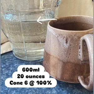 May include: A clear glass measuring pitcher with metric and imperial markings filled with liquid, positioned next to a brown ceramic mug. Text on the image reads 600ml, 20 ounces, and Cone 6 @ 100%. A white arrow points from the pitcher to the mug.