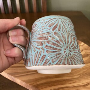 May include: A ceramic mug featuring a light blue glaze over a textured, reddish-brown clay body. The surface is decorated with a repeating carved floral pattern. The mug has a sturdy handle and a flat, unglazed white base with a raised foot ring.