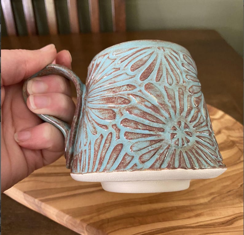 May include: A ceramic mug featuring a light blue glaze over a textured, reddish-brown clay body. The surface is decorated with a repeating carved floral pattern. The mug has a sturdy handle and a flat, unglazed white base with a raised foot ring.