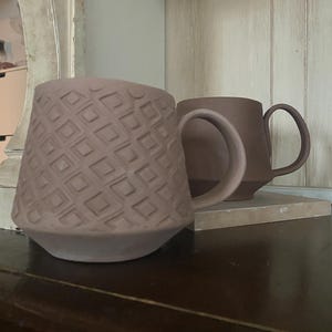 May include: Two matte mauve ceramic mugs with handles. The front mug features a raised geometric pattern of repeating squares across its exterior surface, while the second mug positioned behind it has a smooth, plain finish in the same color.