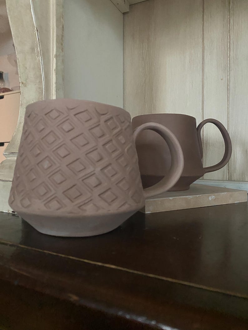 May include: Two matte mauve ceramic mugs with handles. The front mug features a raised geometric pattern of repeating squares across its exterior surface, while the second mug positioned behind it has a smooth, plain finish in the same color.