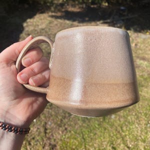May include: A ceramic mug with a tapered, wide-bottomed shape and a curved handle. The mug features a two-tone glaze, with a lighter, speckled beige finish on the upper portion and a darker, solid tan glaze on the lower section of the body.
