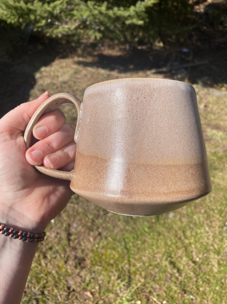 May include: A ceramic mug with a tapered, wide-bottomed shape and a curved handle. The mug features a two-tone glaze, with a lighter, speckled beige finish on the upper portion and a darker, solid tan glaze on the lower section of the body.
