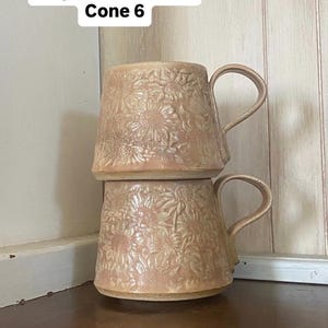 May include: Two stacked ceramic mugs with a light beige, textured sunflower relief pattern and a glossy finish. The text "Glaze: Mayco sand dollar Cone 6" is visible at the top. Each mug features a curved handle and a wide, tapered base design.