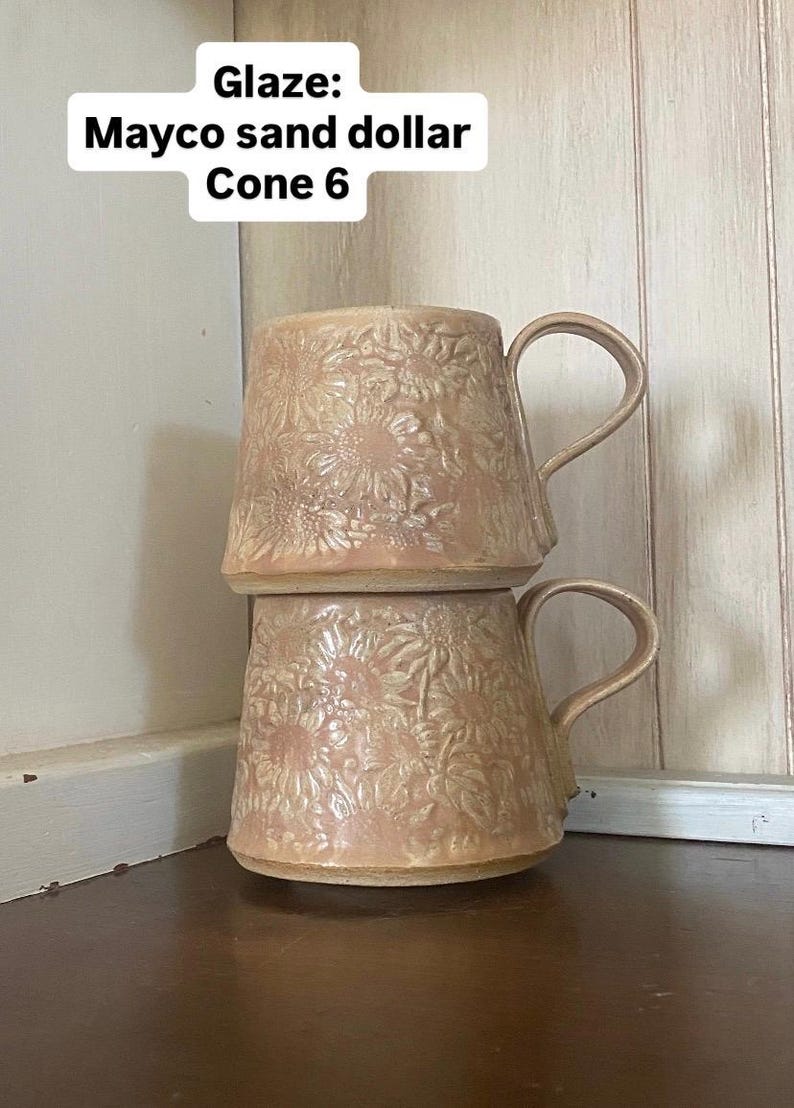 May include: Two stacked ceramic mugs with a light beige, textured sunflower relief pattern and a glossy finish. The text "Glaze: Mayco sand dollar Cone 6" is visible at the top. Each mug features a curved handle and a wide, tapered base design.
