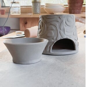 May include: A gray ceramic oil warmer with a floral design and a small gray ceramic bowl. The oil warmer has an opening at the bottom. Other pottery items are visible in the background, including bowls and pots.
