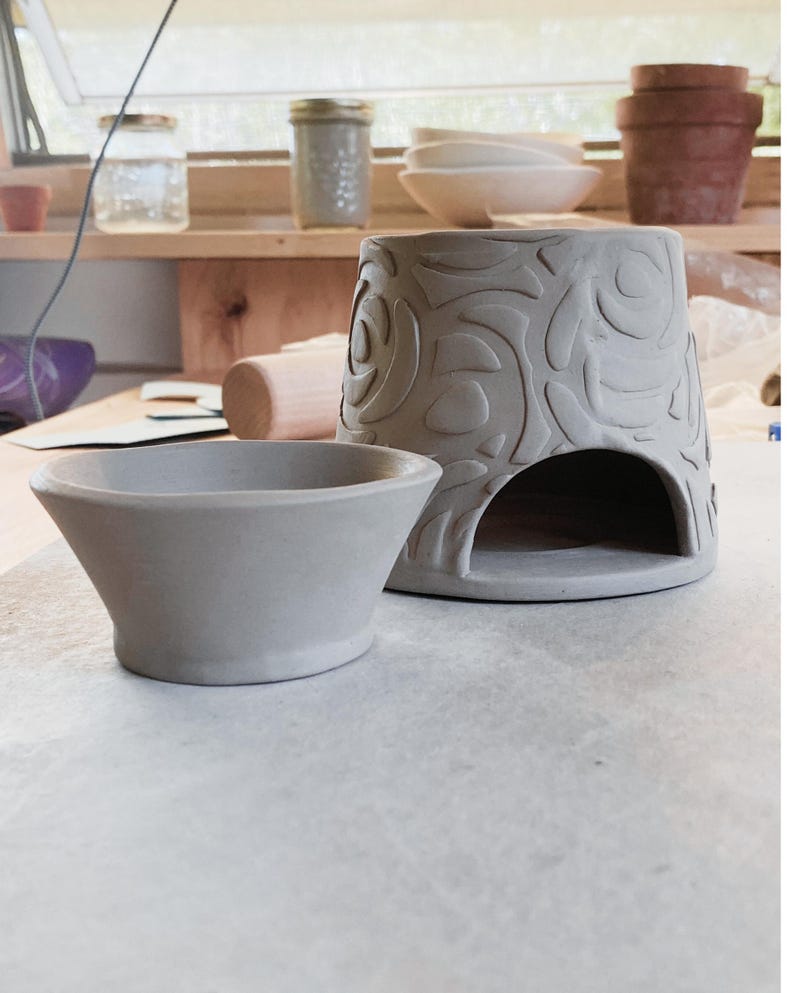 May include: A gray ceramic oil warmer with a floral design and a small gray ceramic bowl. The oil warmer has an opening at the bottom. Other pottery items are visible in the background, including bowls and pots.