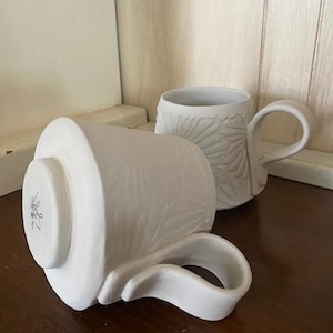 May include: Two white ceramic mugs featuring a raised floral relief pattern on the exterior. One mug stands upright, while the other rests on its side, revealing a circular base with a small, dark, handwritten signature in the center of the bottom.