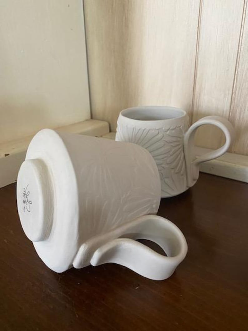 May include: Two white ceramic mugs featuring a raised floral relief pattern on the exterior. One mug stands upright, while the other rests on its side, revealing a circular base with a small, dark, handwritten signature in the center of the bottom.