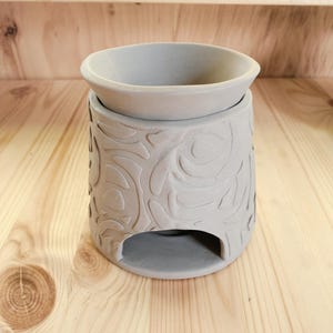 May include: A gray ceramic essential oil warmer with a floral embossed design. The warmer has a round bowl at the top and a rectangular opening at the base. The warmer is sitting on a wooden surface.