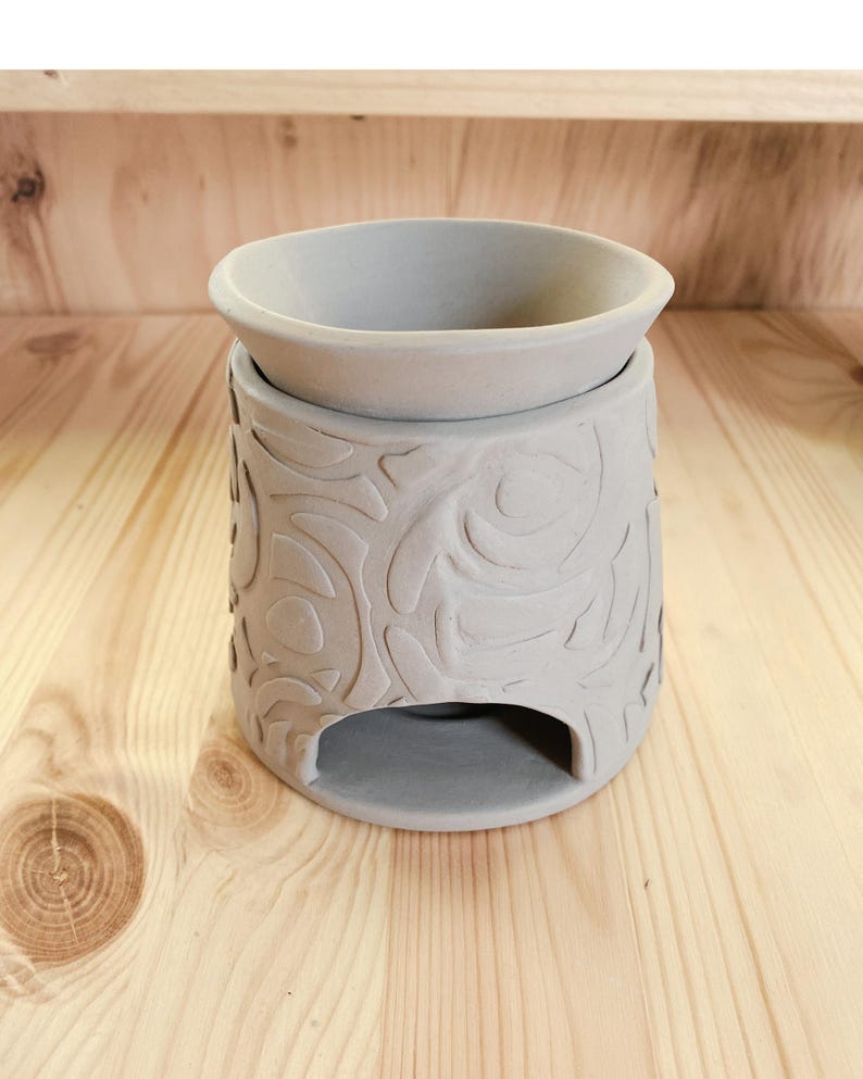 May include: A gray ceramic essential oil warmer with a floral embossed design. The warmer has a round bowl at the top and a rectangular opening at the base. The warmer is sitting on a wooden surface.