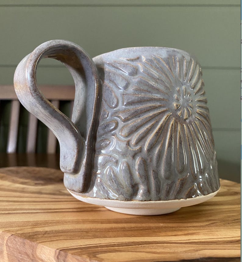 May include: A ceramic mug with a glossy, light grey glaze and a textured, embossed sunburst pattern covering the exterior. The mug features a wide, curved handle and a smooth, unglazed white base that contrasts with the grey body of the vessel.