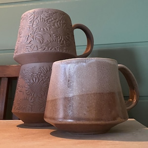 May include: Two ceramic mugs with handles. One mug features a matte brown finish with a raised, embossed sunflower pattern covering the exterior. The second mug has a smooth, two-toned glaze with a lighter beige top and a darker brown base.