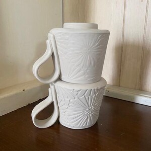 May include: Two stacked white ceramic mugs featuring a matte finish and a relief-carved floral pattern on the exterior. Each mug has a distinctively shaped, curved handle extending from the side, with a tapered body and a flat rim.