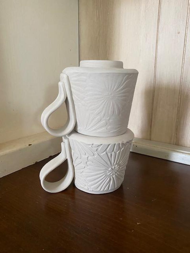 May include: Two stacked white ceramic mugs featuring a matte finish and a relief-carved floral pattern on the exterior. Each mug has a distinctively shaped, curved handle extending from the side, with a tapered body and a flat rim.