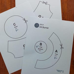 May include: Four white paper templates for an oil burner, featuring dashed lines, scissor icons, and text labels like "Base," "Cup," and "OB 01 Oil Burner." The templates are arranged on a wooden surface.