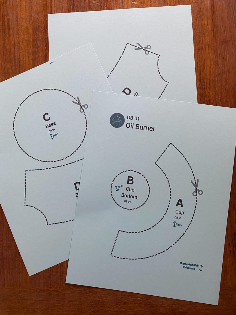 May include: Four white paper templates for an oil burner, featuring dashed lines, scissor icons, and text labels like "Base," "Cup," and "OB 01 Oil Burner." The templates are arranged on a wooden surface.