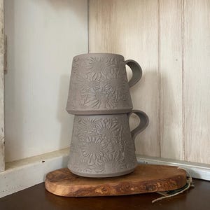 May include: Two stacked matte taupe ceramic mugs featuring a raised, embossed sunflower pattern across the exterior surface. Each mug has a curved handle extending from the side, and the design displays a consistent floral texture throughout.
