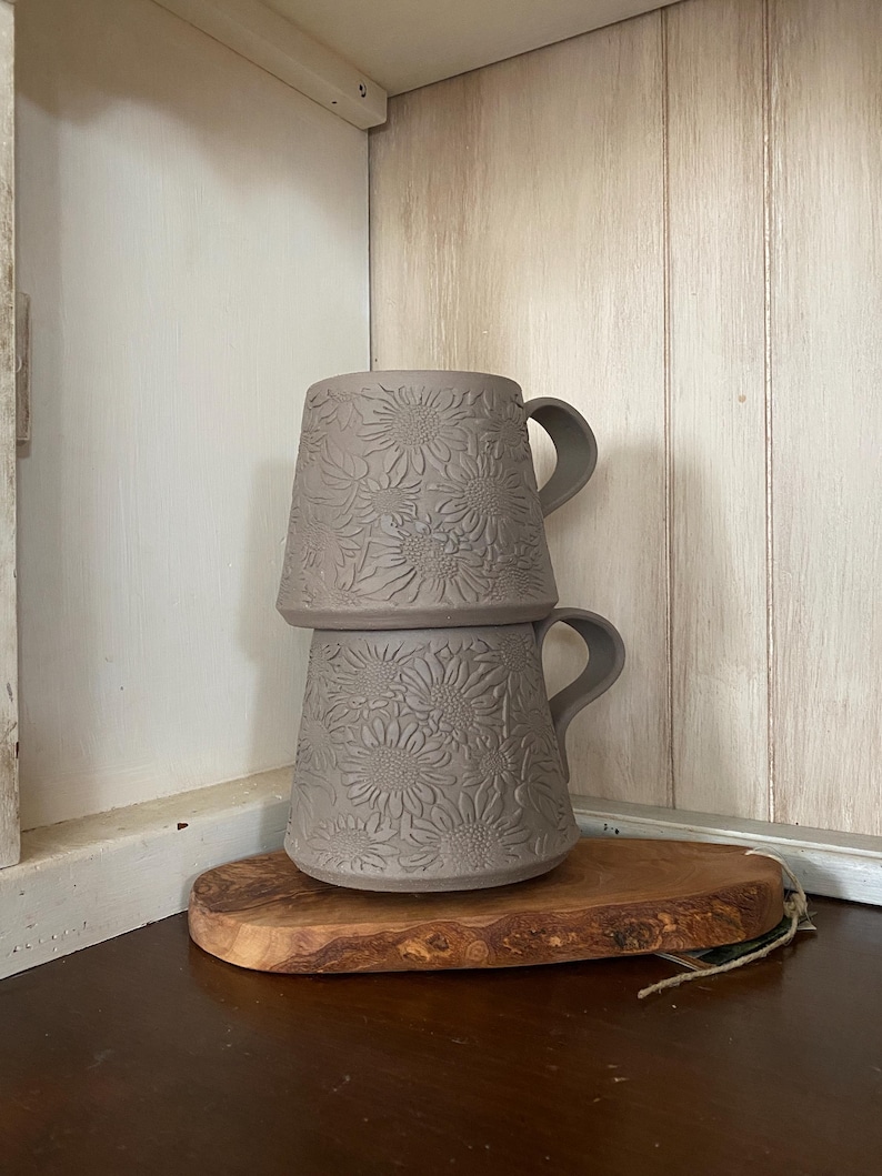 May include: Two stacked matte taupe ceramic mugs featuring a raised, embossed sunflower pattern across the exterior surface. Each mug has a curved handle extending from the side, and the design displays a consistent floral texture throughout.