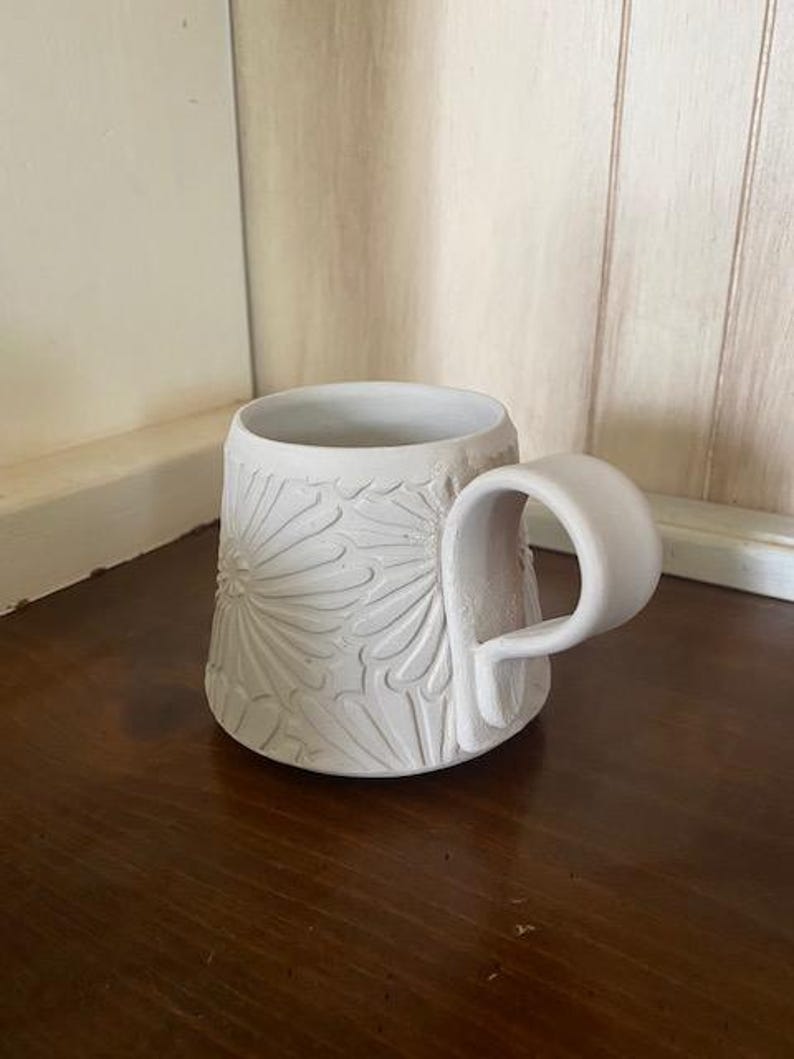 May include: A matte white ceramic mug featuring a tapered silhouette and a wide, curved handle. The exterior surface is decorated with a repeating embossed floral pattern that covers the body of the mug, creating a textured, tactile finish.