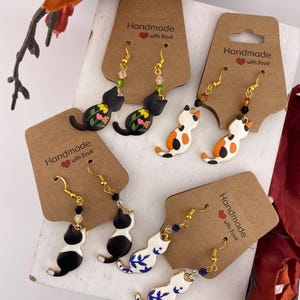 Könnte beinhalten: Handgefertigte Katzenohrringe in verschiedenen Designs. Die Ohrringe zeigen schwarze, weiße und orangefarbene Katzen mit floralen und gemusterten Details. Jedes Paar ist an einer braunen Karte mit dem Text "Handmade with love" befestigt.