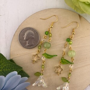 May include: A pair of gold-tone earrings with a floral theme. Each earring features clear and green beads, small green leaves, and clear star-shaped charms. The earrings are displayed on a wooden surface next to a quarter.