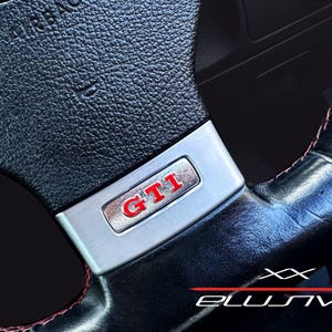 Puede incluir: Primer plano de un volante de cuero negro con costuras rojas. El volante presenta un emblema GTI plateado con letras rojas y un logotipo de Volkswagen. La palabra "AIRBAG" es visible.