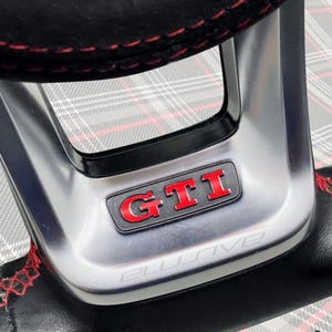 Könnte beinhalten: Nahaufnahme eines schwarzen Lenkrads mit roten Nähten. In der Mitte befindet sich eine silberne Platte mit dem roten "GTI"-Logo und dem Wort "AIRBAG". Das Wort "ELUSIVE" steht unten.