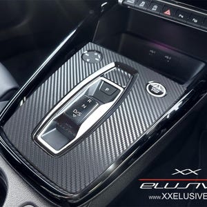 Op de afbeelding: Close-up van de middenconsole van een auto met een koolstofvezelpatroon. De console bevat een versnellingspook, een start/stop-knop en diverse bedieningsknoppen. Het logo "XX Elusive" en het websiteadres zijn zichtbaar.