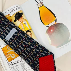Könnte beinhalten: Eine marineblaue Krawatte mit einem sich wiederholenden gold-weißen Kettenmuster und roten Akzenten. Die Krawatte hat ein rotes Futter und ein Etikett mit der Aufschrift "HALSTON". Im Hintergrund ist eine Zeitschrift mit dem Text "LORD & TAYLOR" zu sehen.