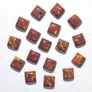 May include: Sixteen square beads with rounded corners, displaying a marbled effect in red, yellow, and brown. The beads are arranged on a white surface, highlighting their unique colour variations. Each bead has a slightly different pattern.