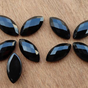 Peut inclure: Huit pierres précieuses noires en forme de marquise avec des surfaces à facettes sont disposées sur une surface en bois. Les pierres précieuses ont un aspect brillant et réfléchissant, et les facettes créent un jeu d'ombre et de lumière.