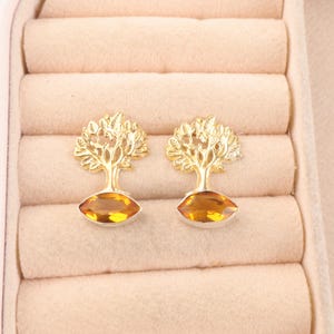 May include: A pair of gold-toned stud earrings featuring a tree design with detailed branches and leaves. Each earring has a marquise-cut amber-coloured gemstone set at the base of the tree trunk, creating a cohesive and symmetrical appearance.