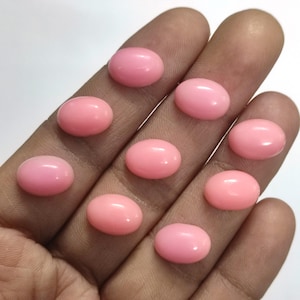 Puede incluir: Colección de nueve piedras preciosas ovaladas de color rosa claro. Las piedras pulidas son lisas y de color uniforme, con un brillo sutil. Las piedras preciosas se muestran en una mano, mostrando su tamaño y forma.