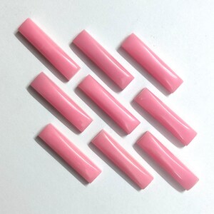May include: Nine rectangular, light pink beads are arranged diagonally on a white surface. Each bead has a slightly rounded shape and a glossy finish. The beads are evenly spaced, creating a visually appealing pattern. These beads could be used for jewelry making or other craft projects.