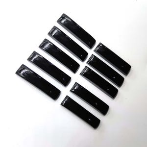 Pode incluir: Uma coleção de dez objetos pretos retangulares brilhantes dispostos diagonalmente sobre uma superfície branca. Cada objeto tem um topo ligeiramente curvo e um acabamento liso e reflexivo. Os itens parecem ser pequenos componentes, possivelmente decorativos ou funcionais.