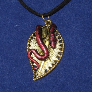 May include: A gold leaf pendant necklace with a red snake design. The snake is curled around the leaf and has a shiny, iridescent finish.