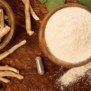 Może przedstawiać: Zdjęcie z g&oacute;ry produkt&oacute;w z ashwagandhy. Dwie drewniane miski zawierają korzeń i proszek ashwagandhy. Kapsułka leży w pobliżu. Przedmioty znajdują się na ciemnej drewnianej powierzchni. Widoczne są zielone liście.