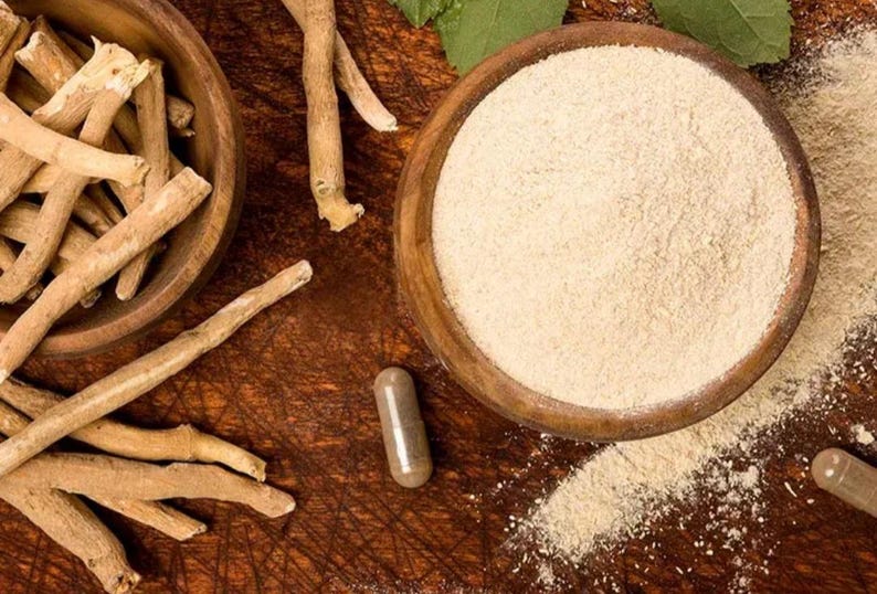 Może przedstawiać: Zdjęcie z g&oacute;ry produkt&oacute;w z ashwagandhy. Dwie drewniane miski zawierają korzeń i proszek ashwagandhy. Kapsułka leży w pobliżu. Przedmioty znajdują się na ciemnej drewnianej powierzchni. Widoczne są zielone liście.