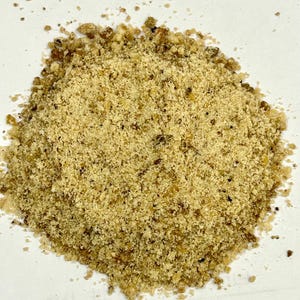 May include: A close-up shot of a pile of finely ground, light brown food product. The texture appears granular, with small, darker flecks interspersed throughout. The food item is likely a spice or seasoning.
