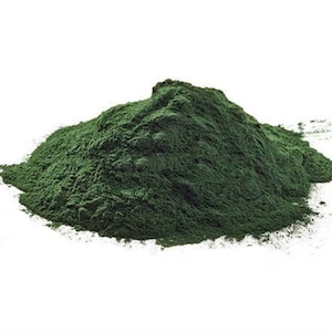May include: A pile of dark green powder against a white background. The powder has a fine, granular texture and appears to be a natural product. The image is well-lit, highlighting the color and texture of the powder.