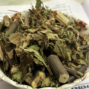 May include: A white bowl filled with dried herbs, leaves, and small wooden pieces. The herbs are a mix of green and brown, with some stems and twigs visible. The bowl has a decorative rim.