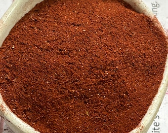 May include: A white ceramic bowl filled with a vibrant red-brown spice. The bowl has a slightly irregular shape and a textured surface. The spice appears finely ground, with a consistent color throughout.
