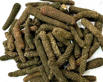 Puede incluir: Un montón de pimientos largos secos, de color verde oscuro y marrón. Las vainas tienen una superficie texturizada y estriada, de forma cilíndrica. La imagen es un primer plano, que muestra los detalles de la especia.